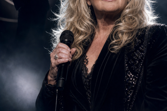 Cantora loira segurando um microfone em um palco, com silhueta de um homem ao fundo e luzes dramáticas